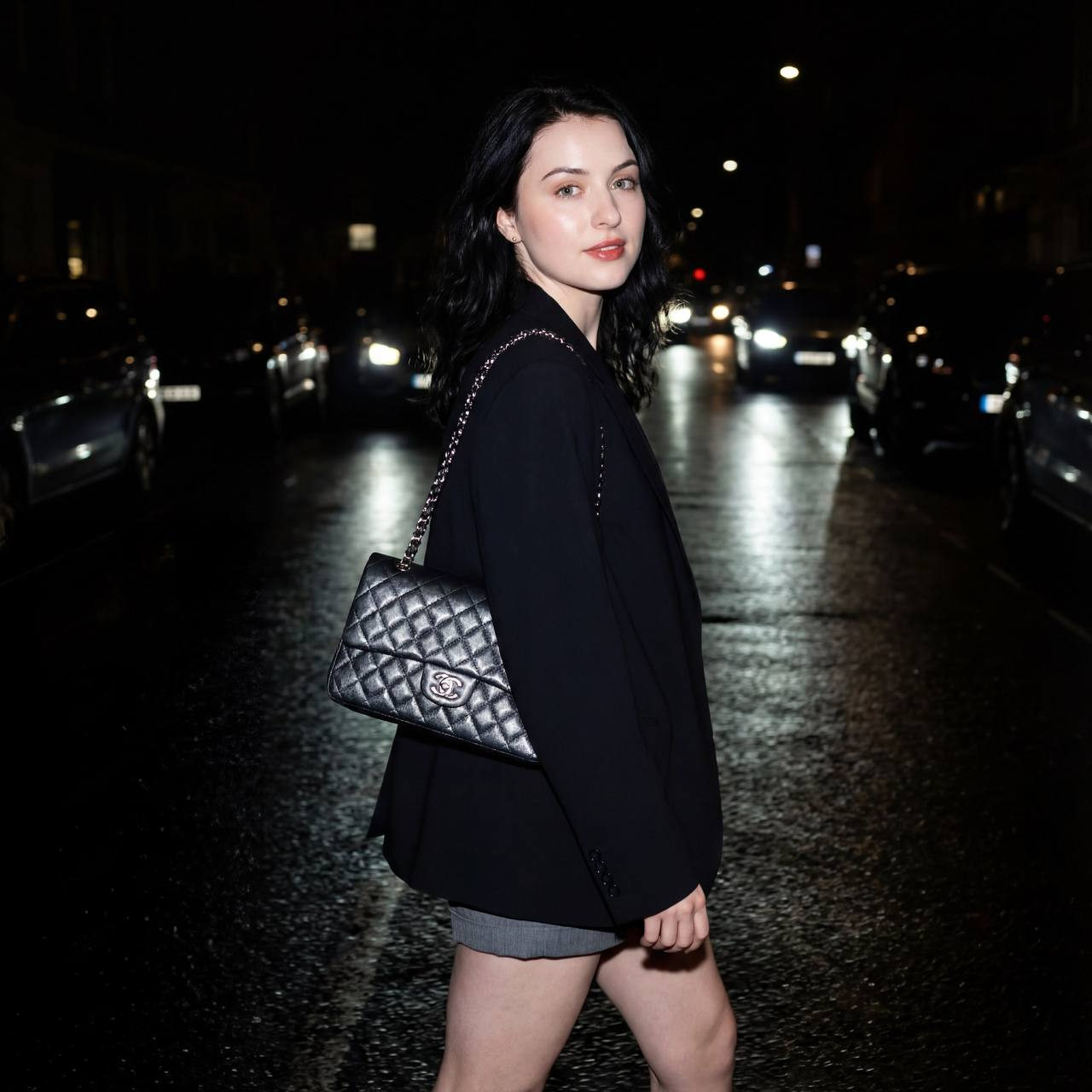 A woman in an oversized blazer and gray shorts poses on a dark street, illuminated by bright flash photography. Her smooth skin and glowing face create a captivating scene.
