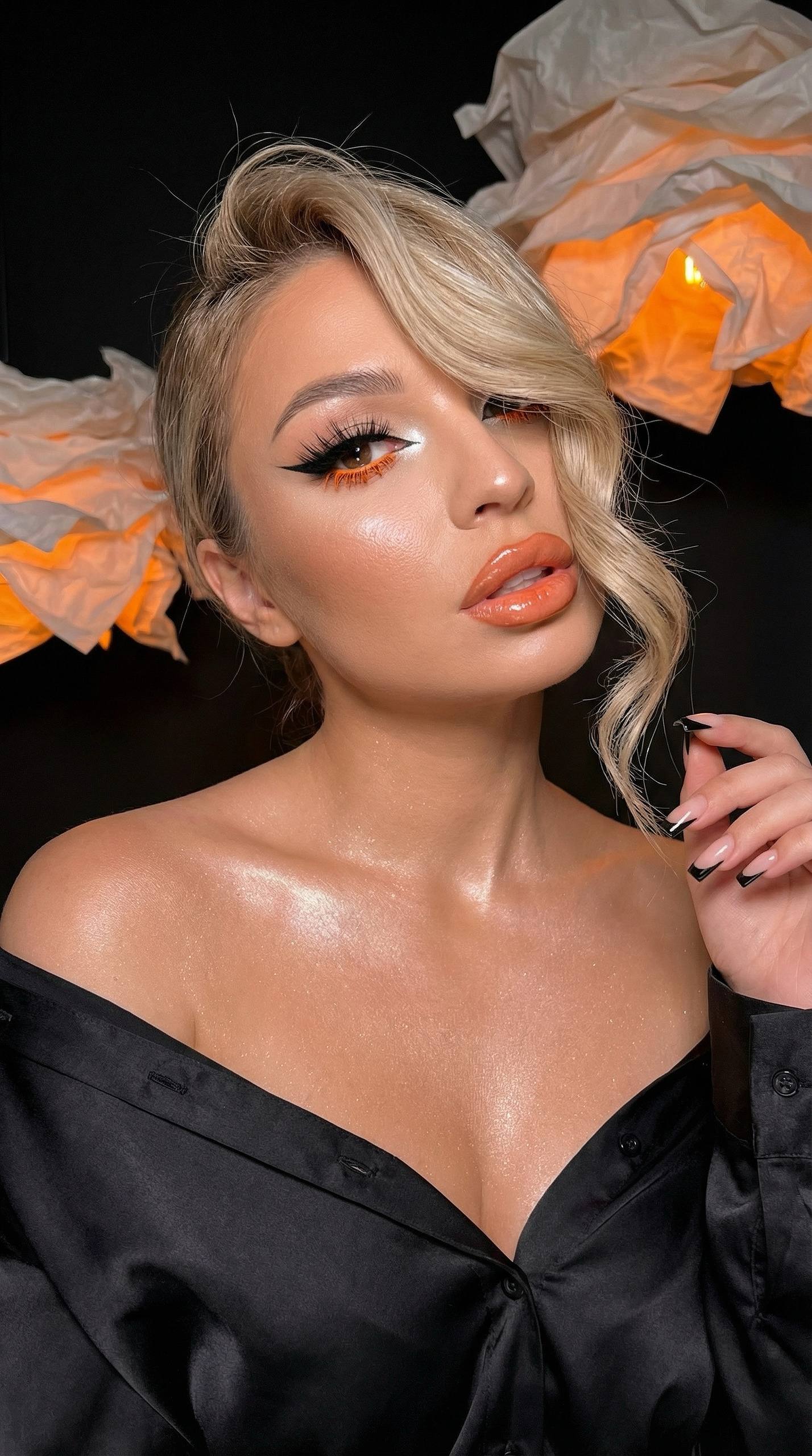 Close-up portrait of a girl with orange makeup, glossy hair, and elegant eye makeup, illuminated against a dark beauty studio.