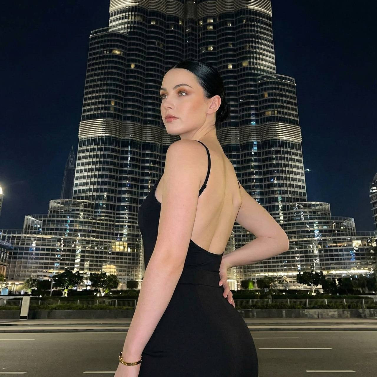 A confident woman in an evening outfit poses on a Dubai street at night, illuminated by a camera flash with skyscrapers in the background.