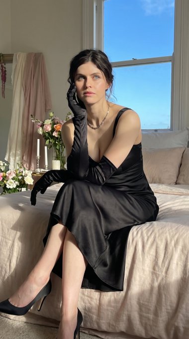 A woman in a black satin slip dress seated on a pale pink bed with bokeh background.