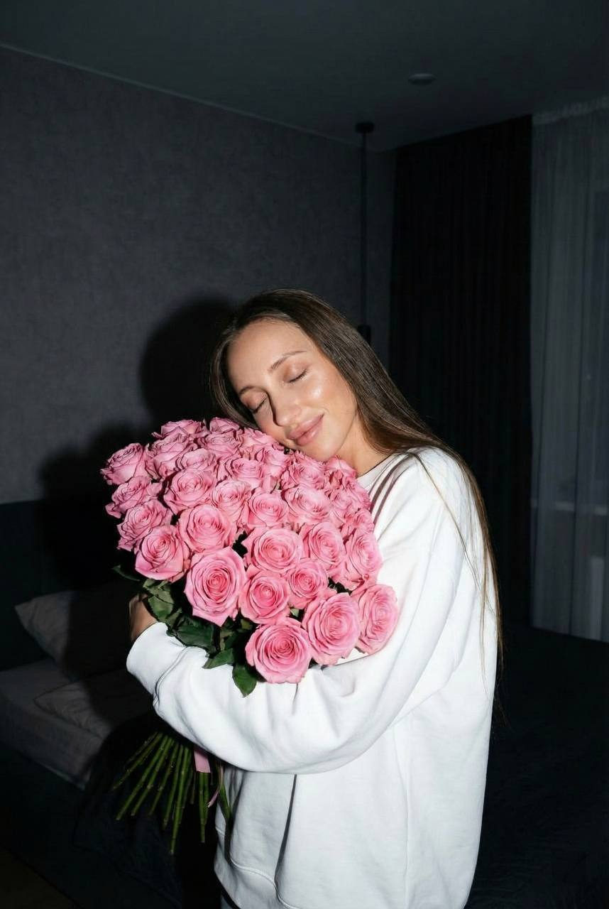 A girl in a white oversized sweatshirt hugs a large bouquet of pink roses, eyes closed, in a dimly lit bedroom.