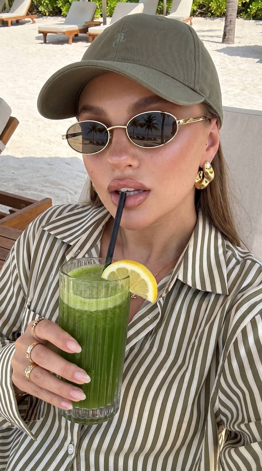 A close-up selfie of a woman with sun-kissed skin at a luxury resort, holding a tall glass of green juice, wearing gold-framed sunglasses.