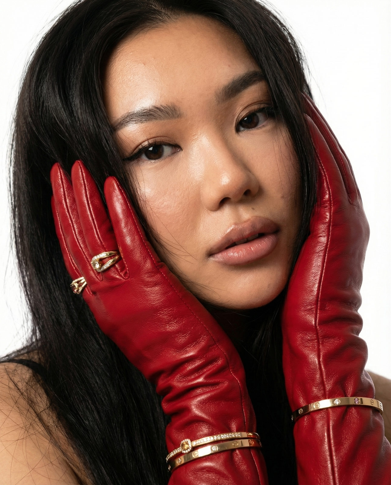 Intimate close-up of a woman softly lit, wearing red leather gloves, showcasing gold rings and a poised expression.
