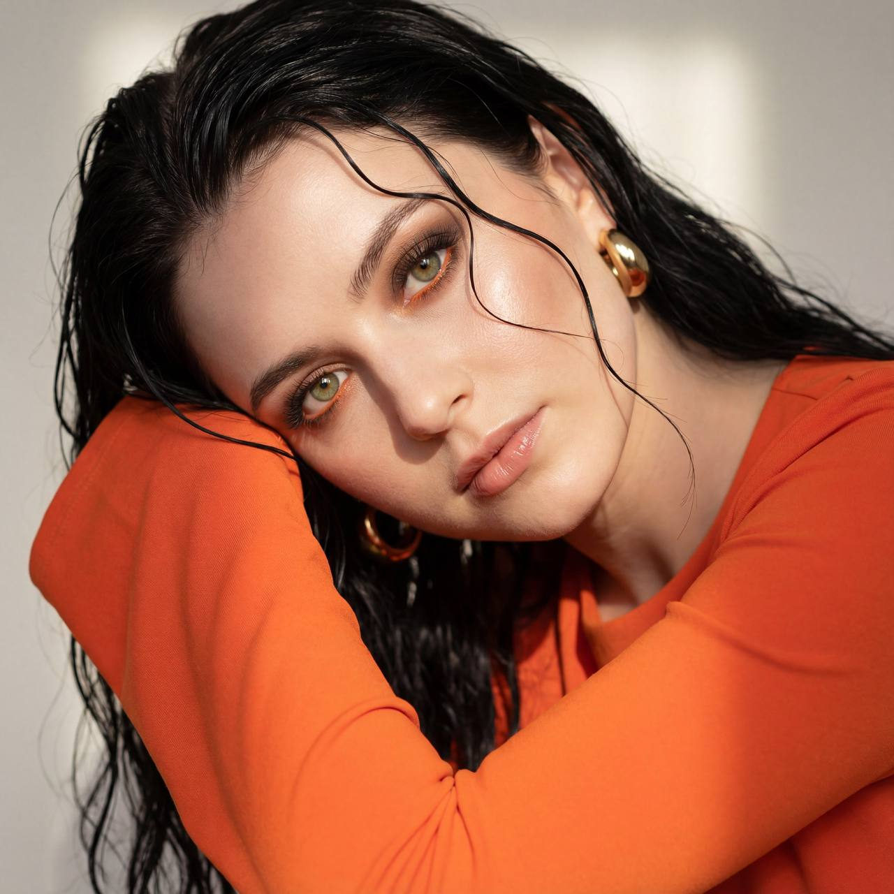 Close-up portrait of a woman with wet hair and a bright orange fitted top, gazing intensely at the camera with soft lighting.