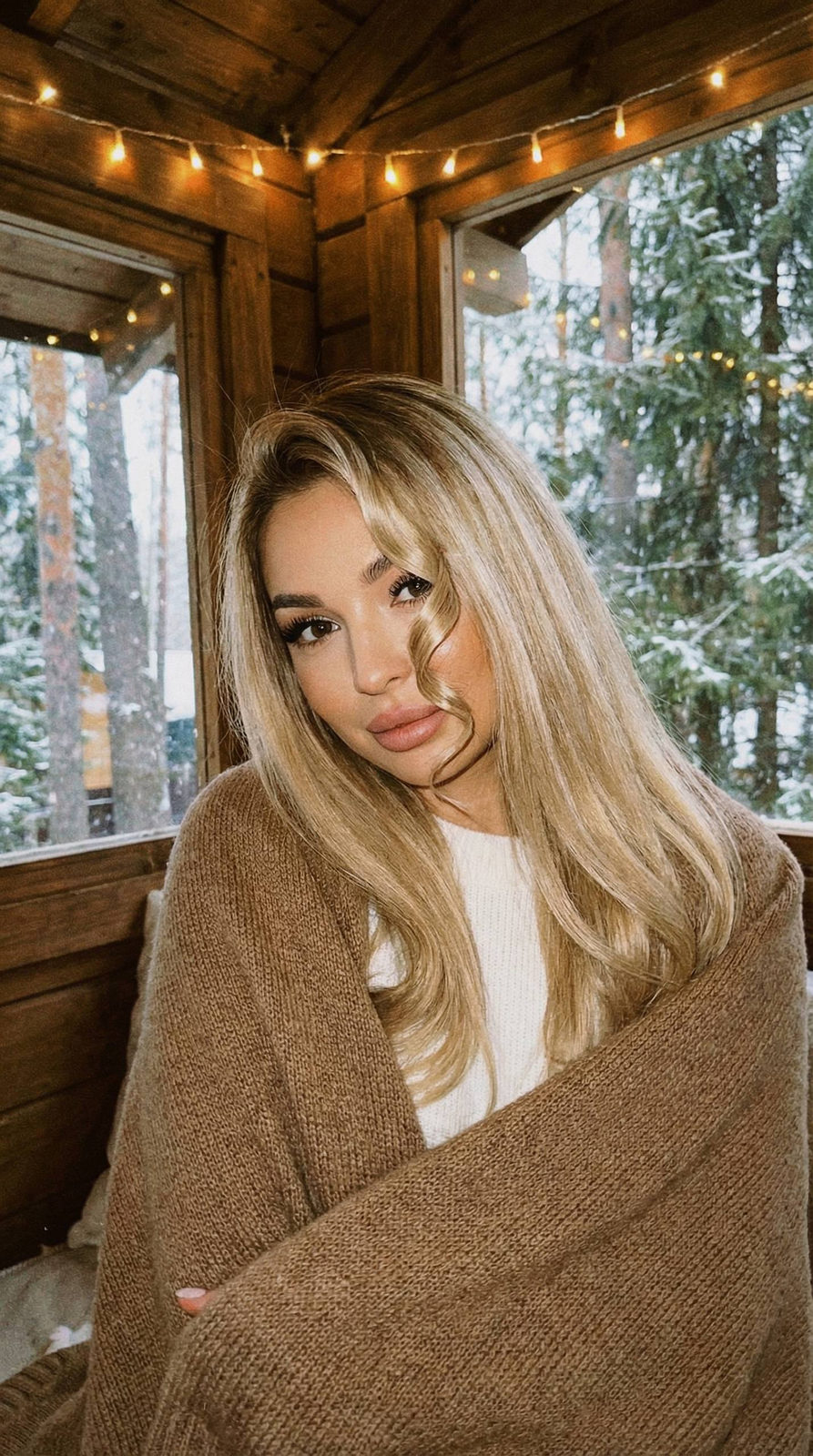A girl with long shiny hair in a cozy treehouse, relaxed in a sweater, as light snow falls around her.