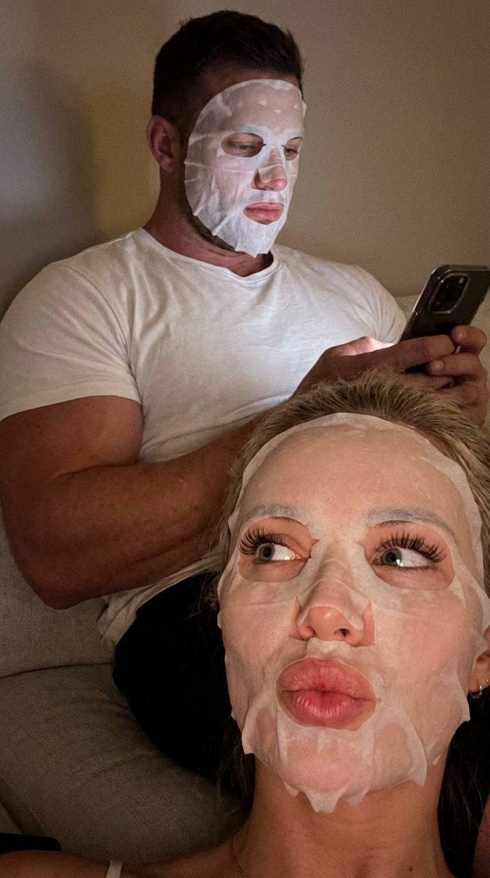 A couple lying comfortably at home, both wearing white cloth face masks in soft, cozy lighting.