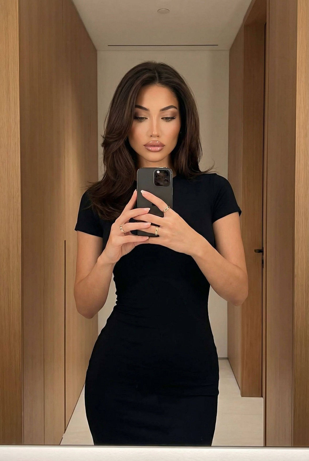 A woman taking a mirror selfie in a modern wood-paneled interior, dressed in a fitted dress and styled hair.