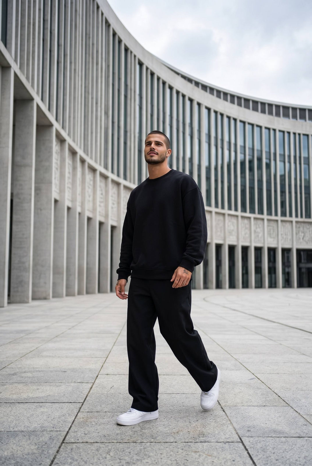 A man in oversized black sweatshirt and trousers walking confidently across a modern plaza before a large curved white building.