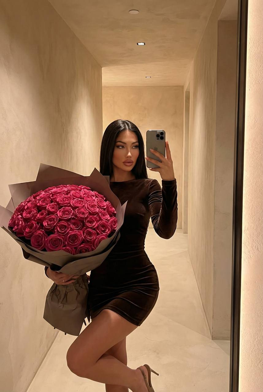 A woman in a dark brown velvet dress stands in a hallway holding a large bouquet of deep pink roses.