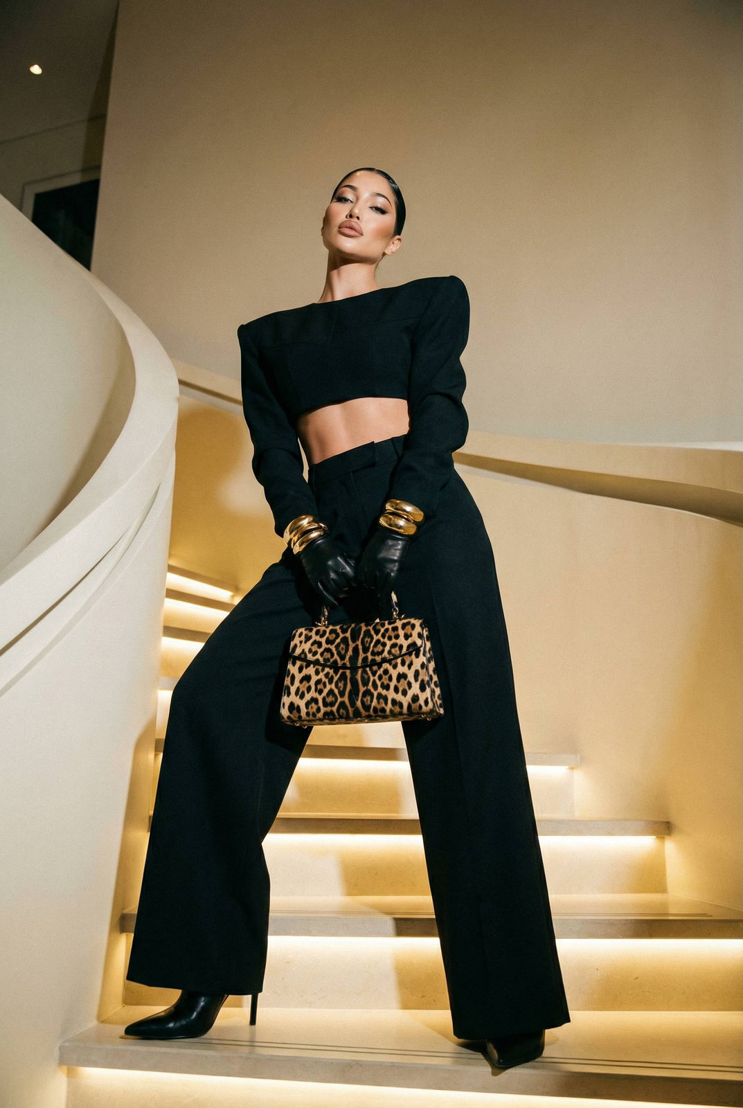 A stylish woman poses on a curved cream staircase wearing a structured crop top and dramatic trousers, illuminated by warm ambient lighting.