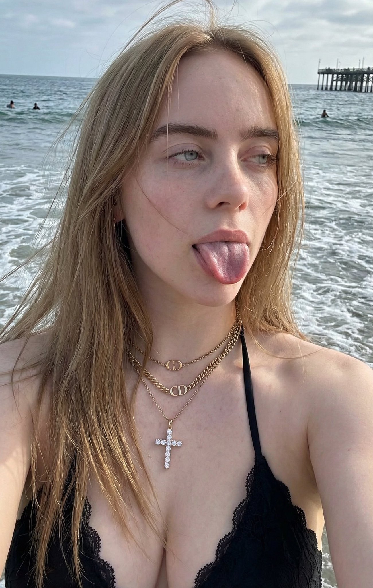 Young woman with fair skin and long light brown hair in a black bikini at the beach, sticking her tongue out playfully.