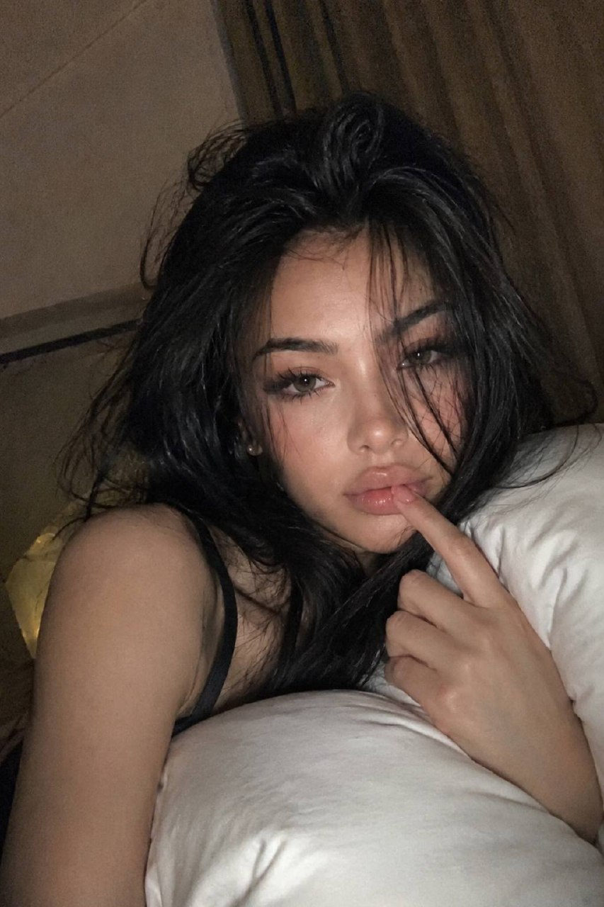 A woman lying on a bed hugging a pillow in a cozy bedroom, with messy long black hair and natural glossy lips, captured in a nostalgic late-night atmosphere.