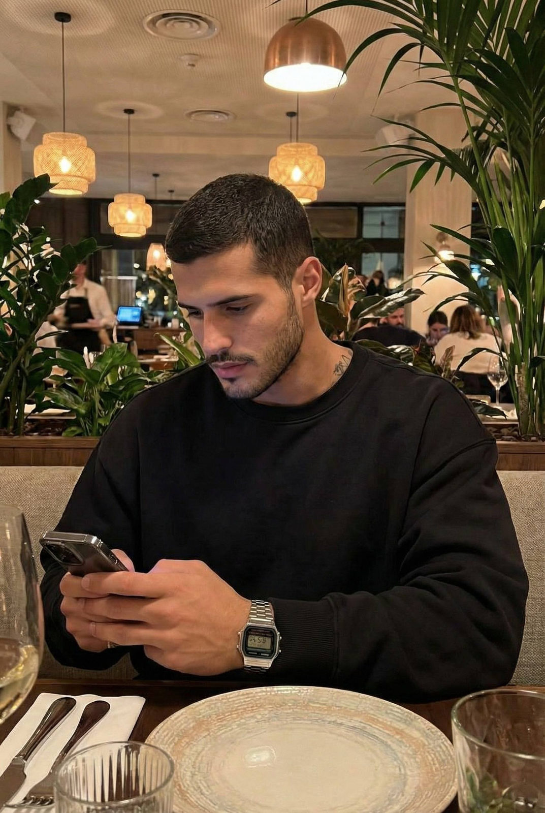 A man sitting at an elegantly set restaurant table with lush plants and warm lighting, immersed in his smartphone.