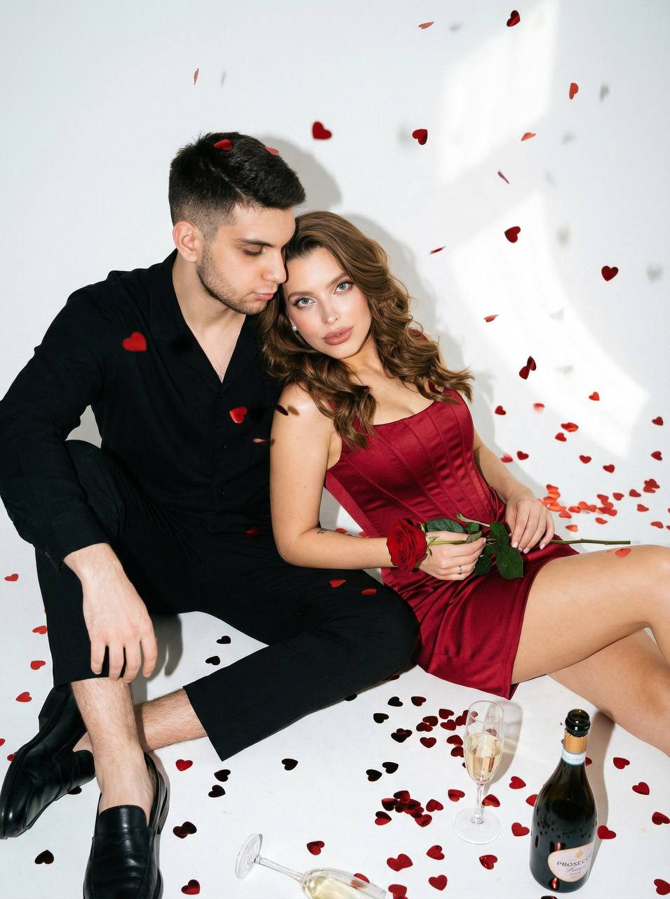 A man and woman sitting on a white floor, surrounded by red heart confetti, both looking engaged and festive.