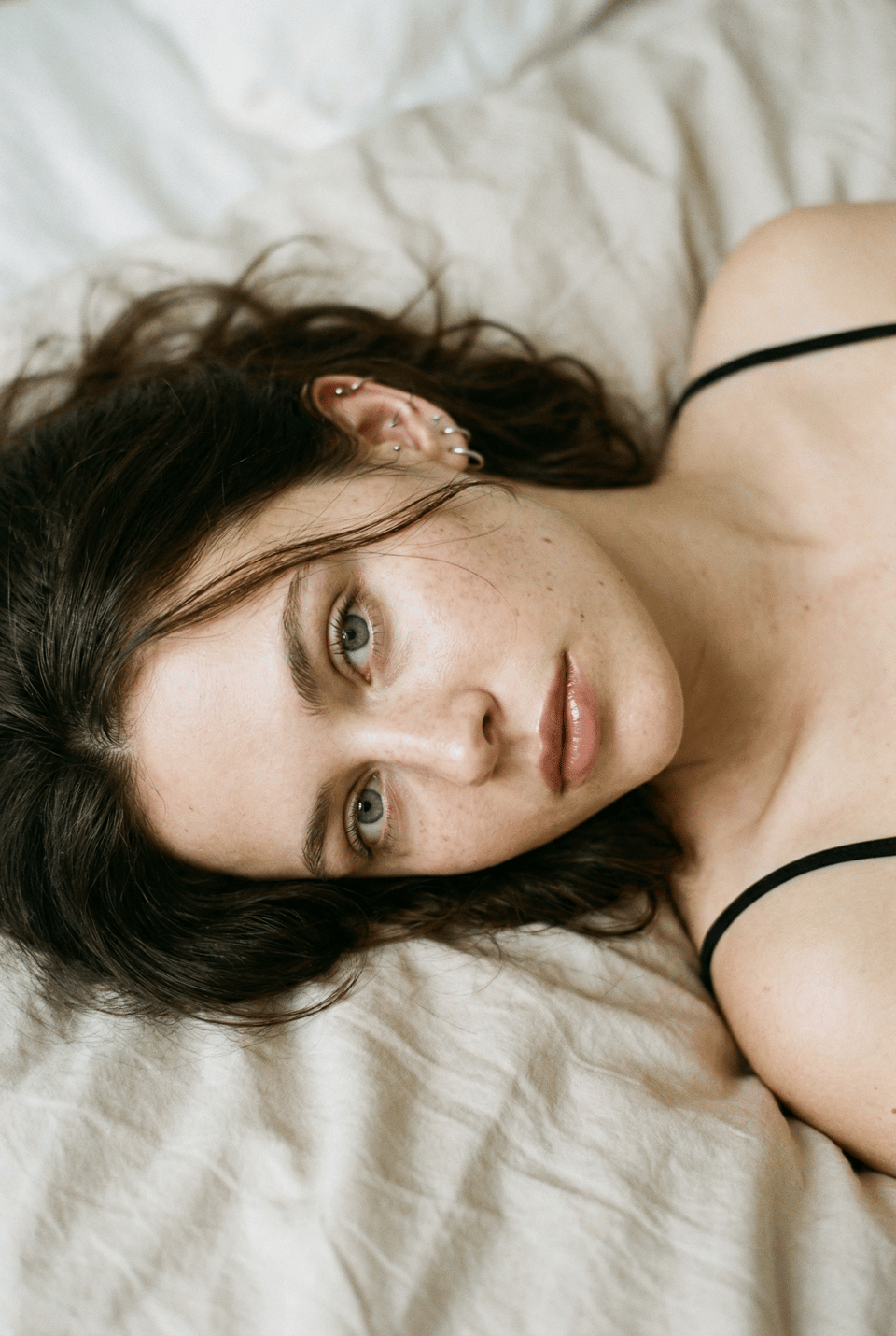 Close-up portrait of a young woman with freckles, relaxed on bed, gazing softly into the camera.