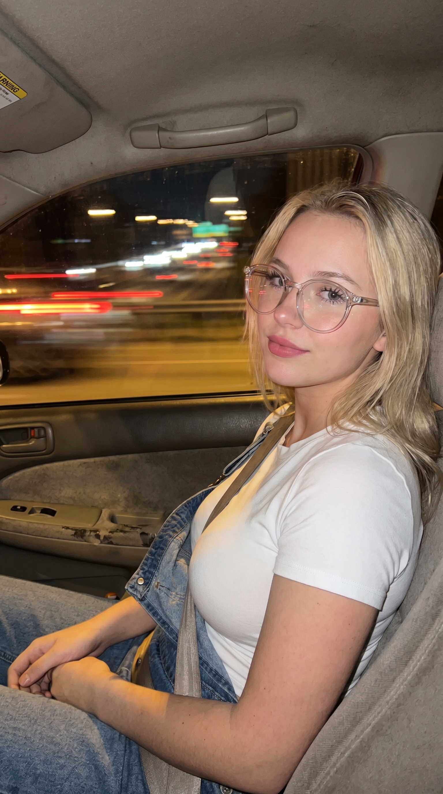 A slim girl with long wavy hair sitting in the passenger seat of a moving car, wearing soft pink lipstick and natural makeup.