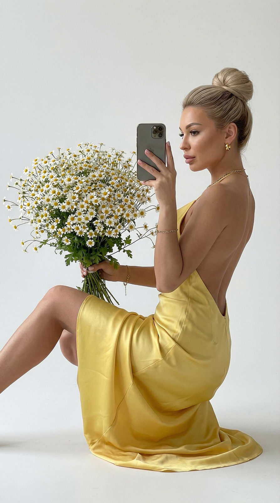 A woman in a backless satin yellow dress sitting gracefully, holding a large bouquet of daisies against a white background.