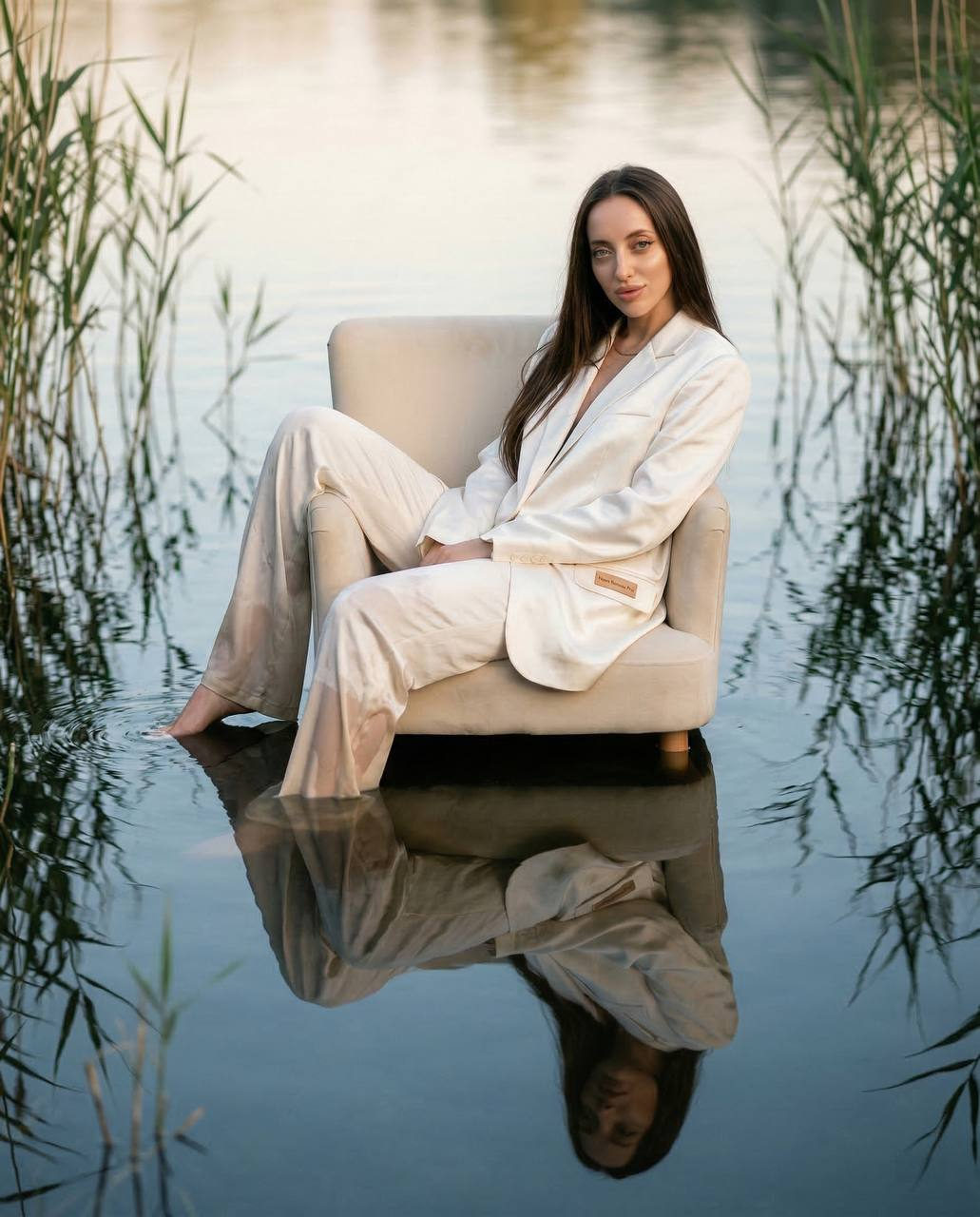 A model in a white oversized silk suit reclines on a cream chair, half immersed in a calm lake, showcasing detailed reflections and soft lighting.