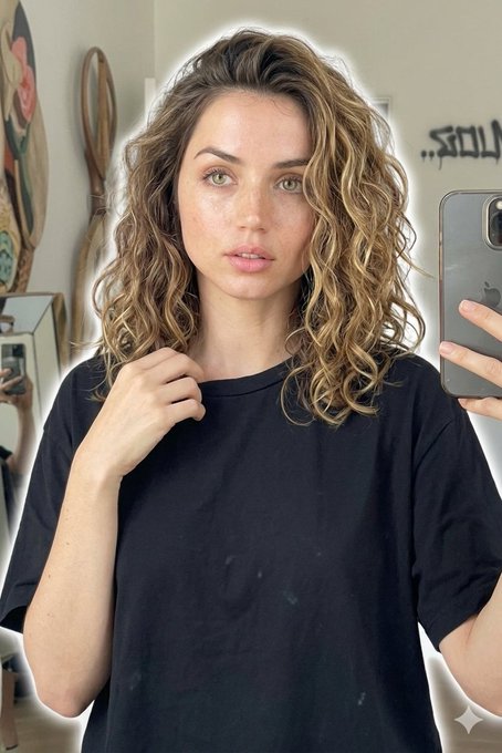 A woman with light brown wavy hair takes a selfie in front of a graffiti wall, holding a smartphone with a soft glow illuminating her features.