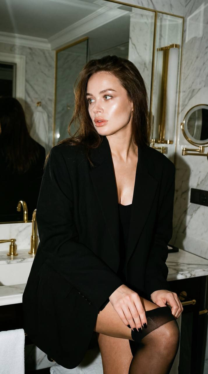 A girl in a luxury hotel bathroom near a mirror, pulling up a black stocking, wearing an oversized black blazer.