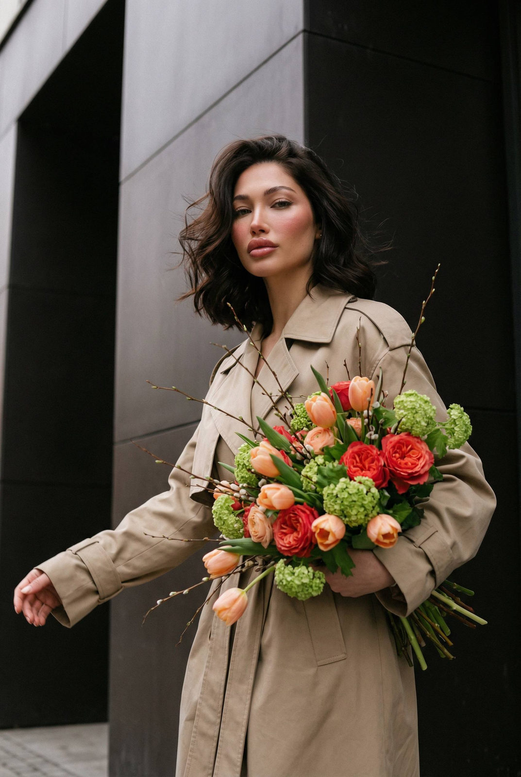 Woman in oversized beige trench coat holding a bouquet of peach tulips and wildflowers in a modern urban setting.