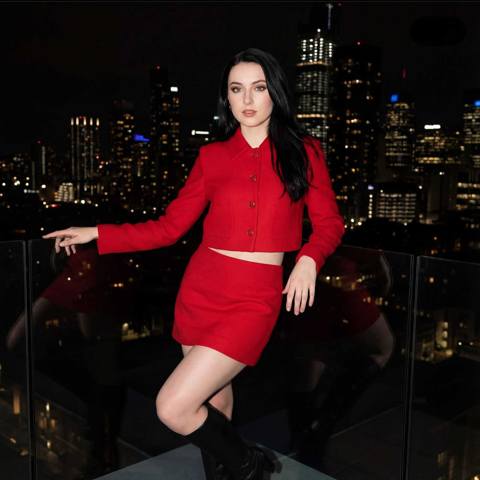A woman in a bright red suit posing on a city rooftop at night, exuding confidence with a stylish cropped jacket and high boots.
