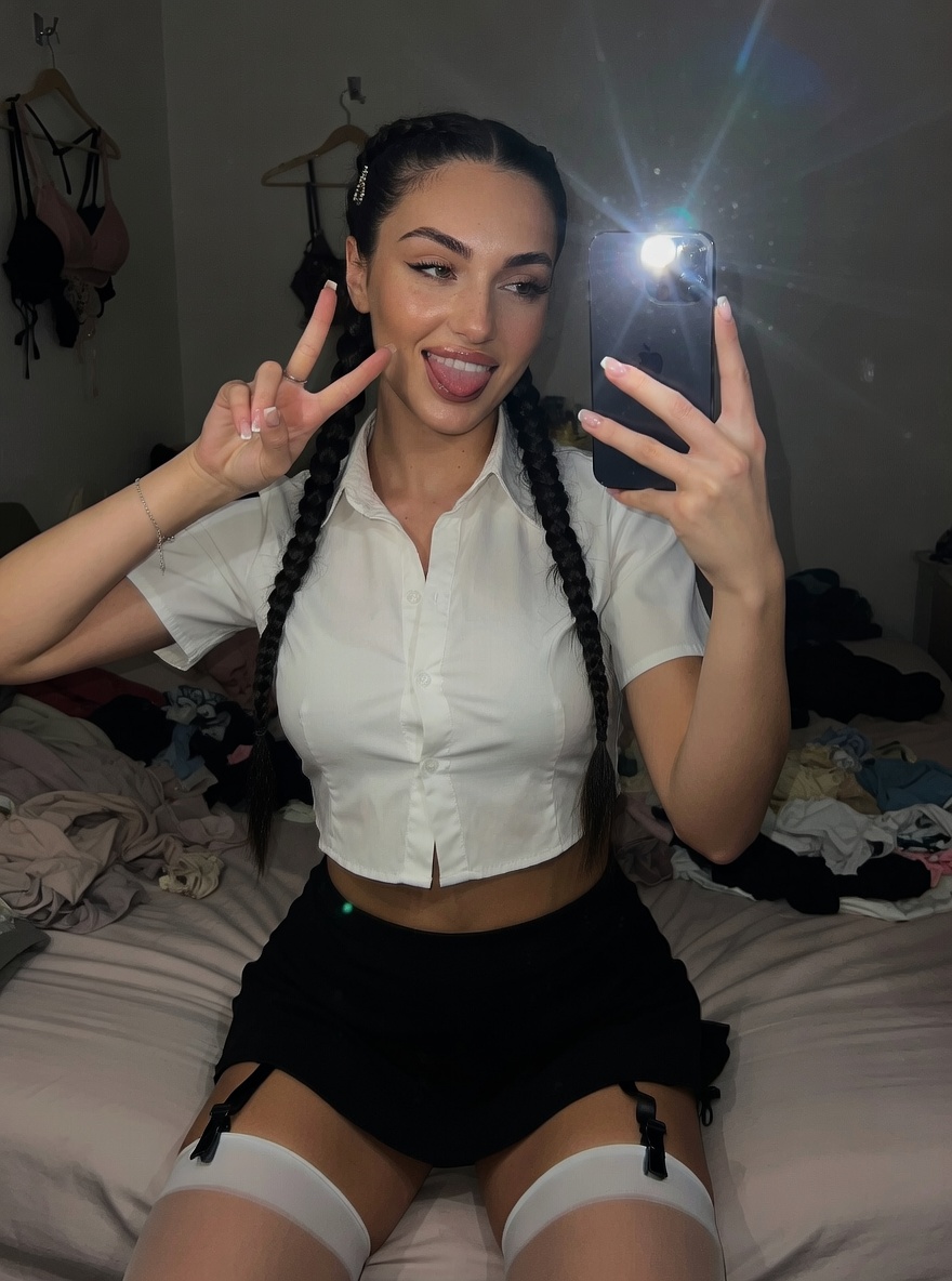 A playful school girl with twin braids posing with a peace sign in a messy bedroom, wearing a cropped white blouse and black mini skirt.