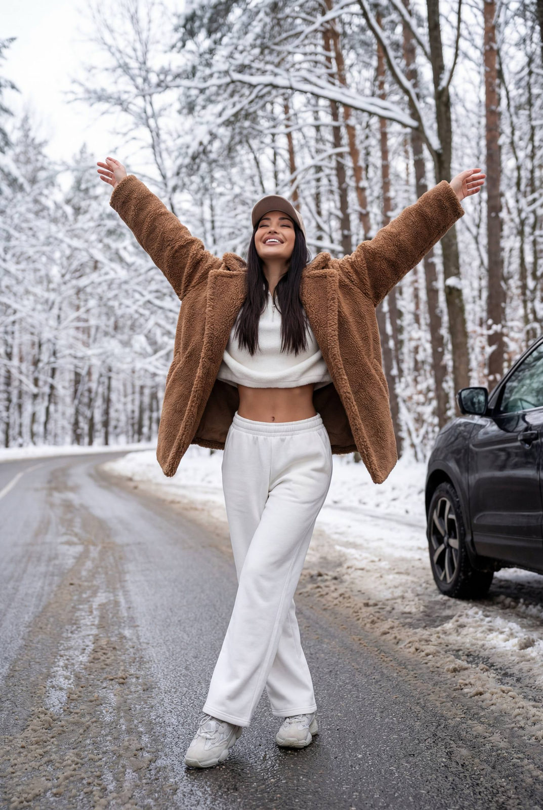 A woman joyfully standing in a snowy forest, wearing an oversized brown teddy jacket and white sweatpants.