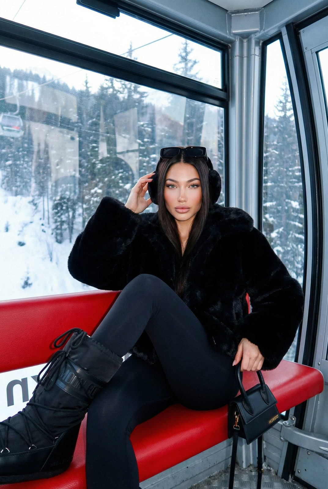 A young woman in luxurious black faux-fur jacket inside a ski lift gondola, adjusting earmuffs against a snowy mountain backdrop.