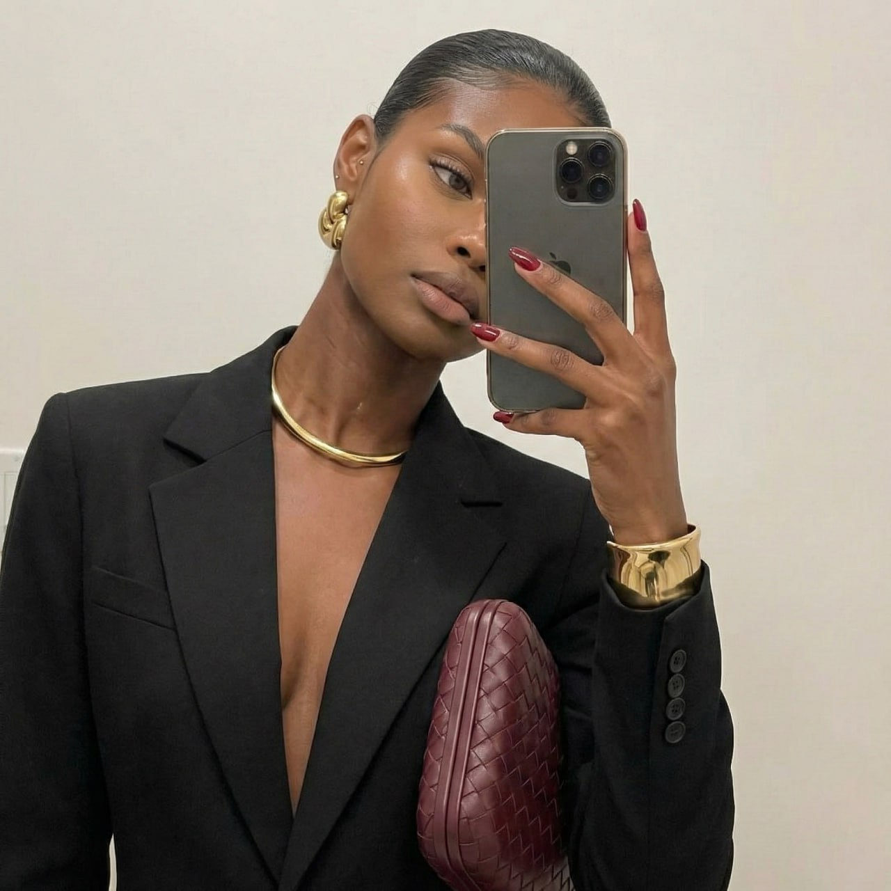 A close-up mirror selfie portrait showcasing a woman in a black blazer with gold accessories, captured in soft ambient light.