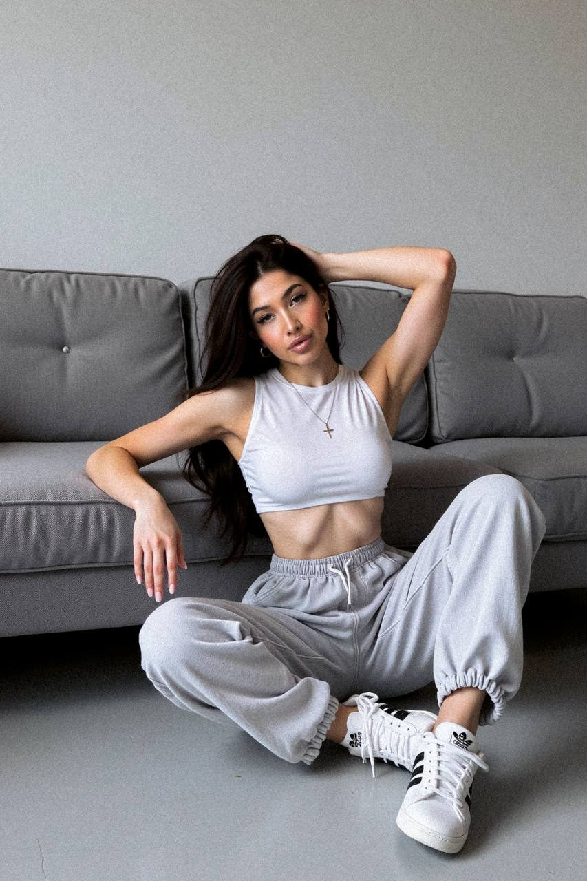 Woman in sporty crop top and oversized grey jeans sitting relaxed on minimalistic sofa, casual pose with natural lighting.