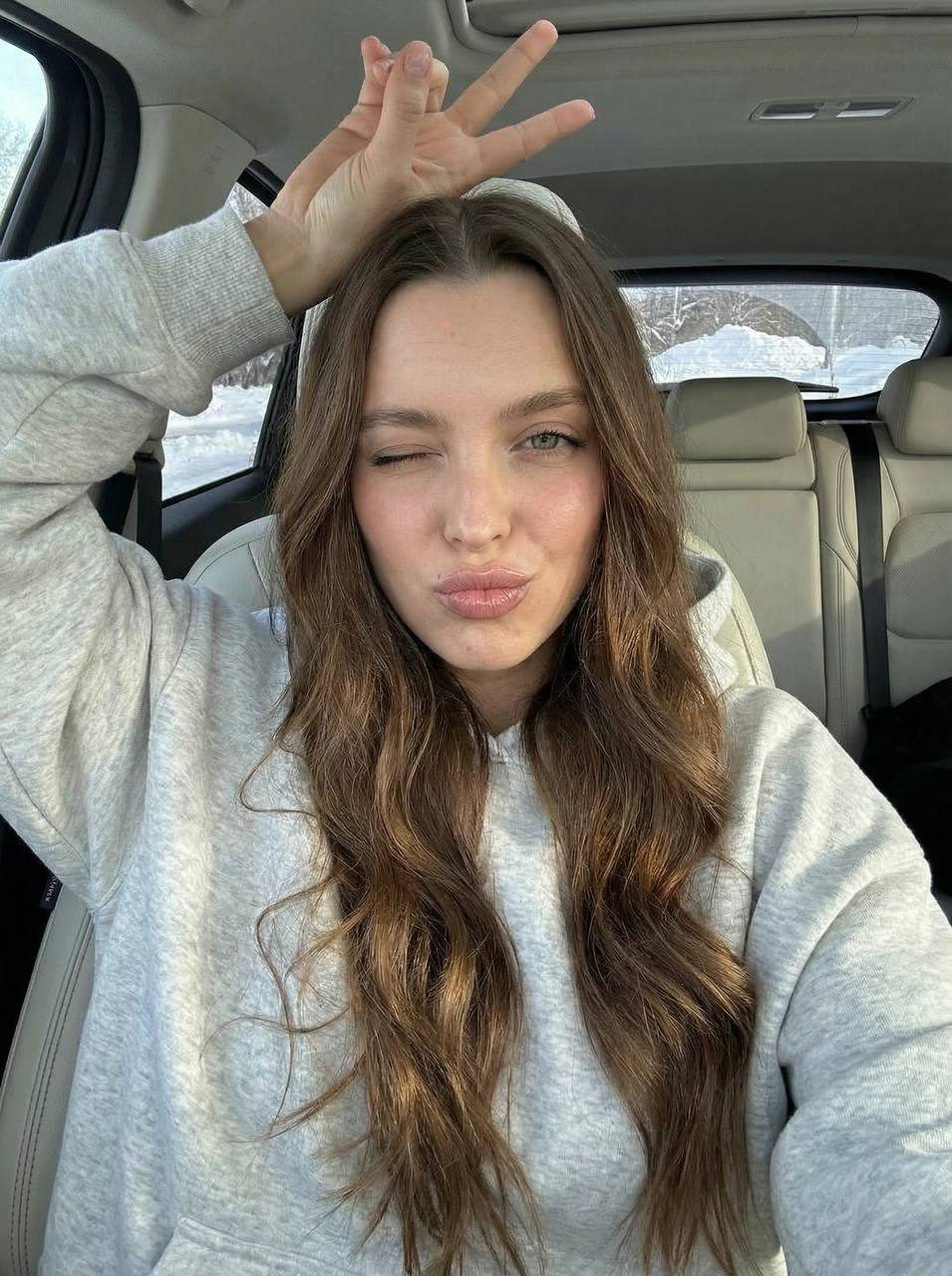 A close-up amateur selfie of a person in a car, winking playfully with fingers raised above the head.
