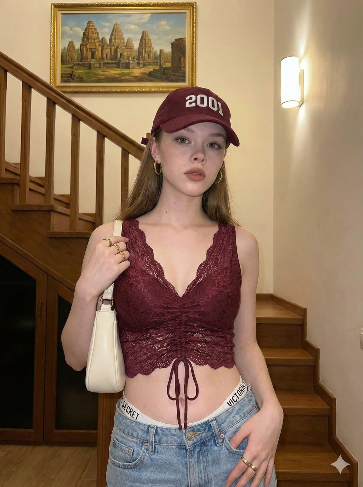 A model in a maroon lace crop top and light blue denim jeans posing indoors with a cozy home setting and golden artwork.