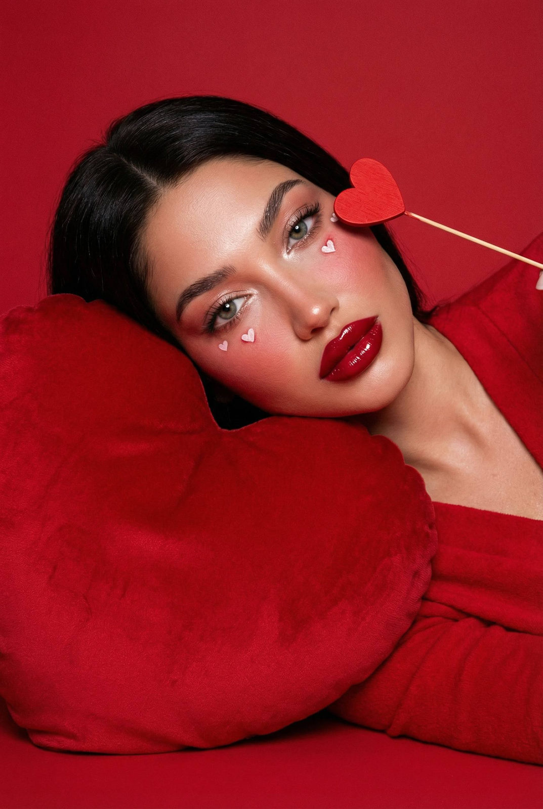 A model in a soft red garment leans against a plush red velvet heart pillow, showcasing rich textures and romantic makeup.