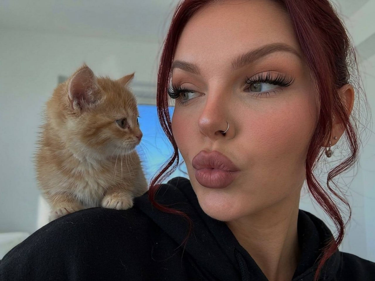 Close-up selfie of a woman with a kitten on her shoulder, lips puckered in a kiss, soft natural lighting.