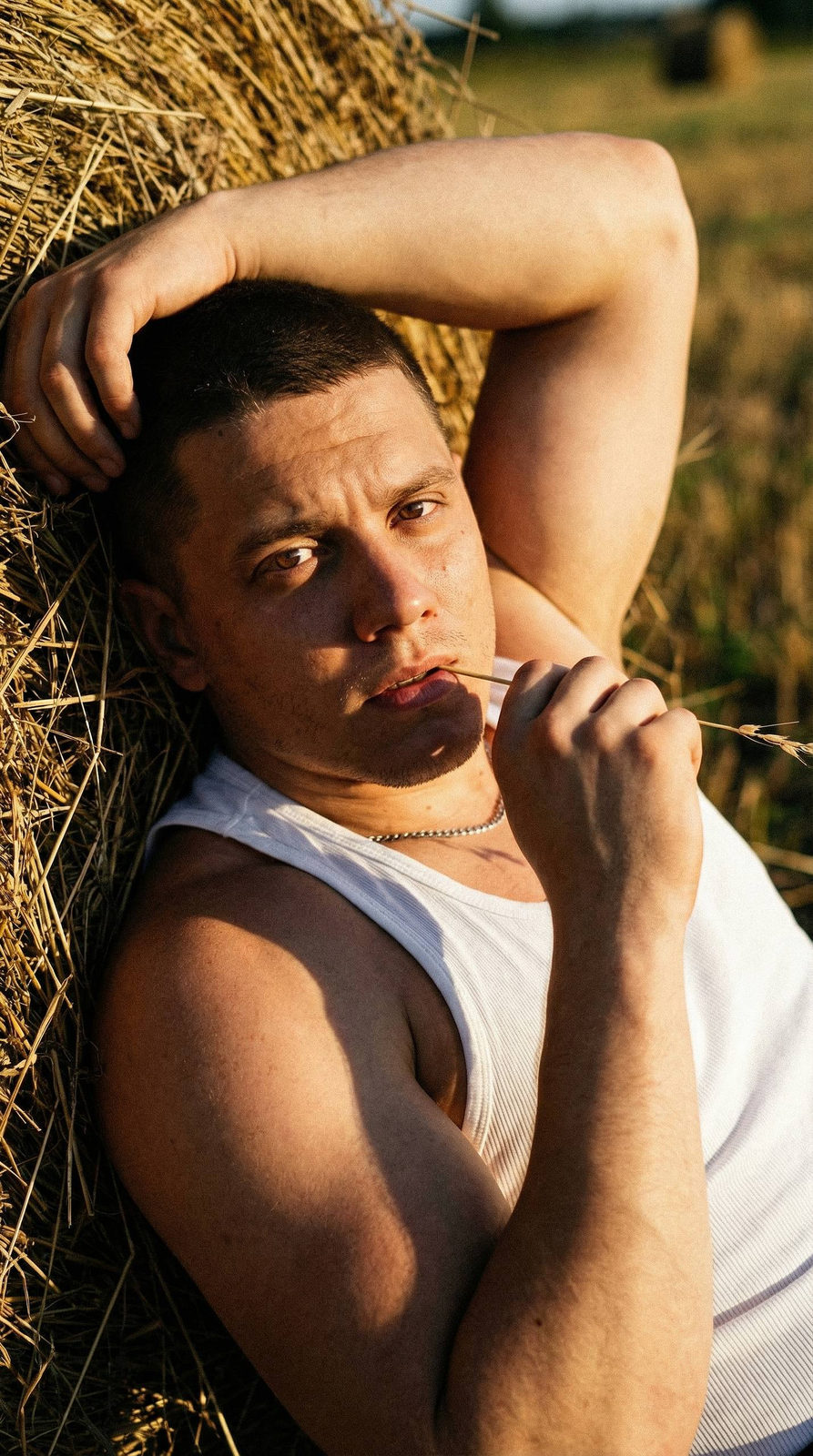 Extreme close-up of a muscular man leaning against a haystack at golden hour, relaxed pose with straw in mouth.
