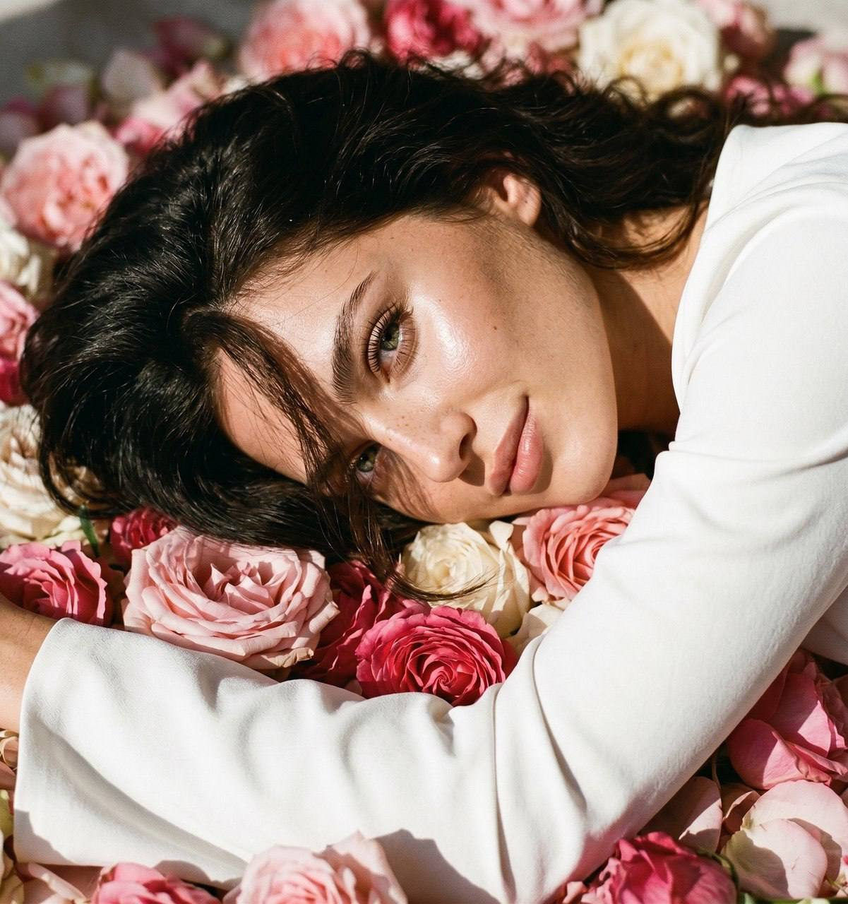 Close-up of a girl with shiny hair posing among pink flowers in a white top.