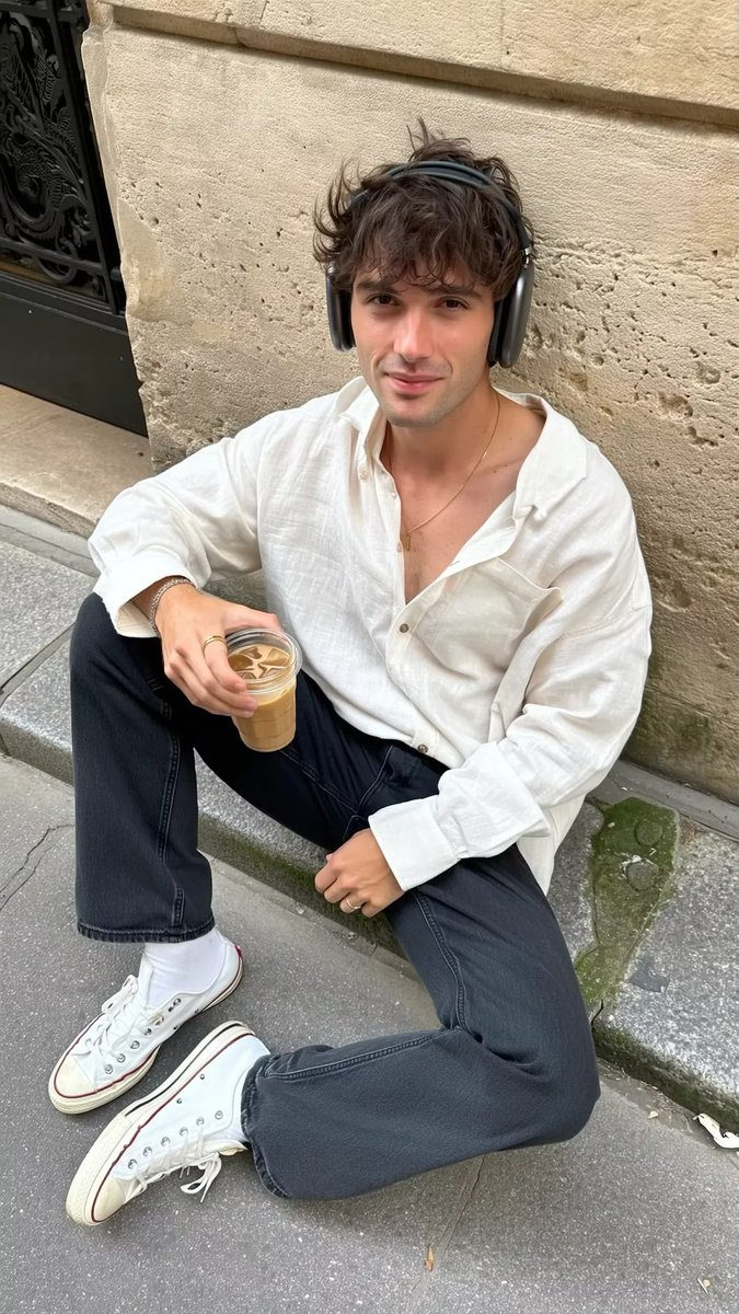 A casually dressed man with tousled dark hair lounging against a weathered Parisian stone wall, holding a translucent iced coffee glass.