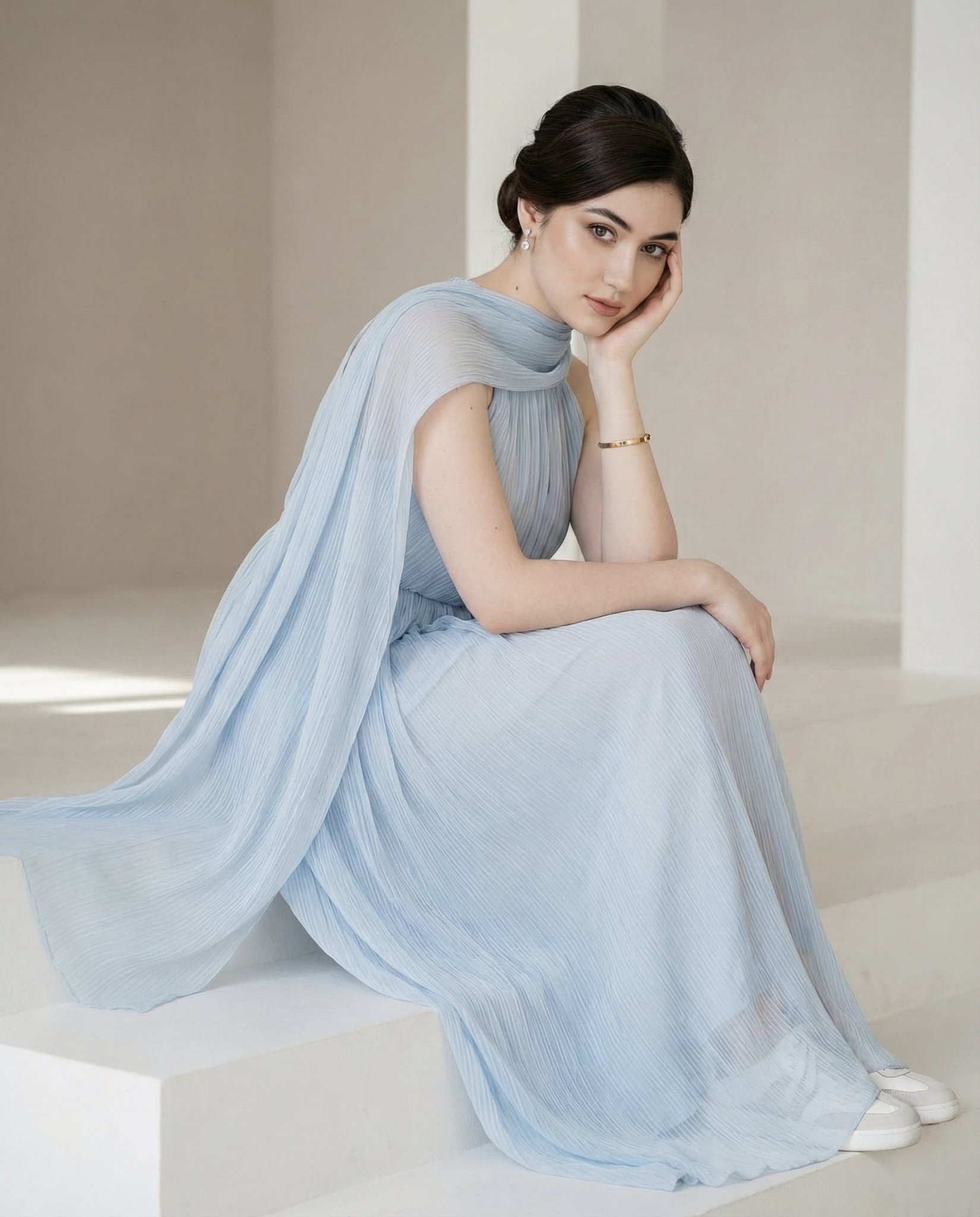 A woman in a flowing pale blue gown sits gracefully on white steps, soft lighting enhancing her calm expression.