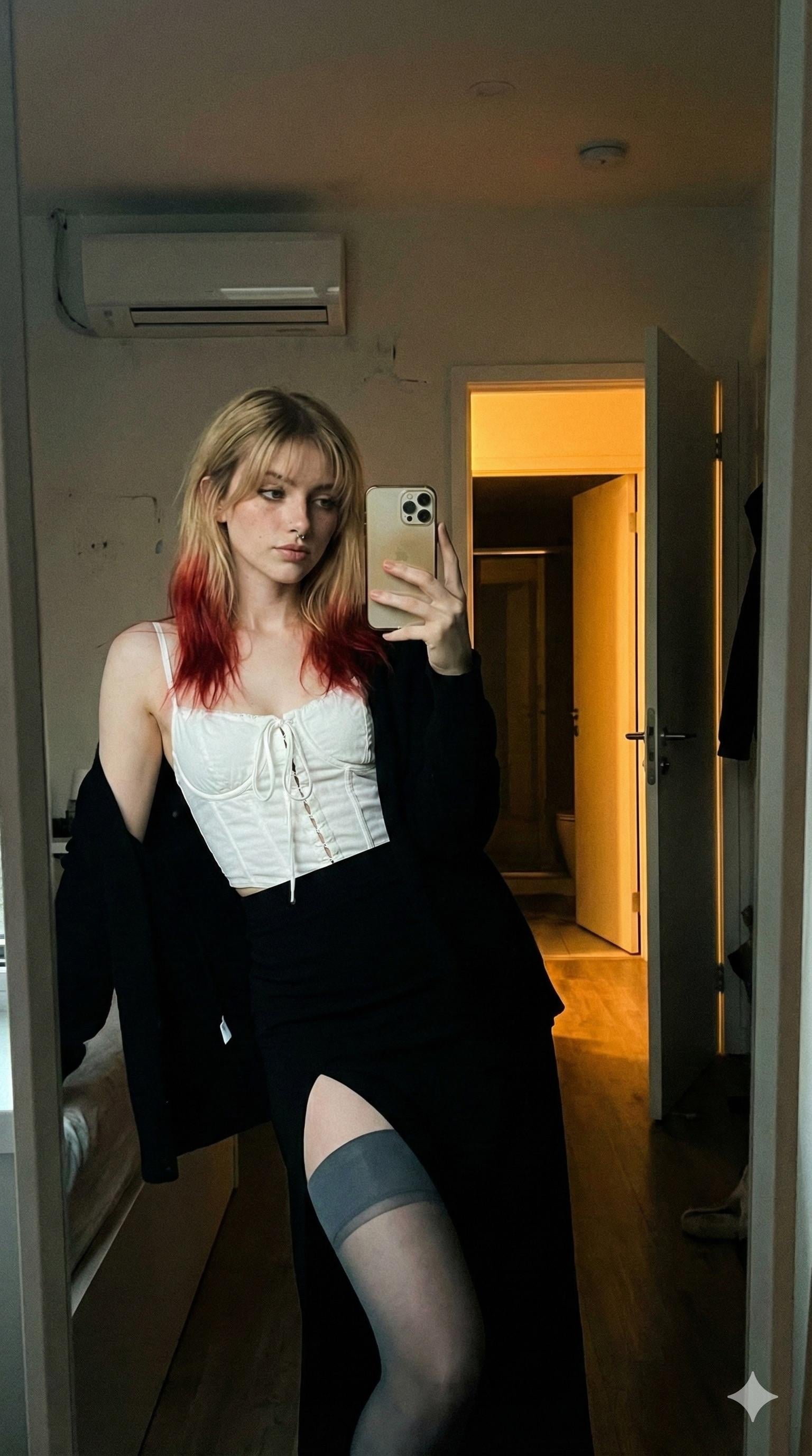A stylish person taking a mirror selfie in a dimly lit room, dressed in a white corset and black high-slit skirt.