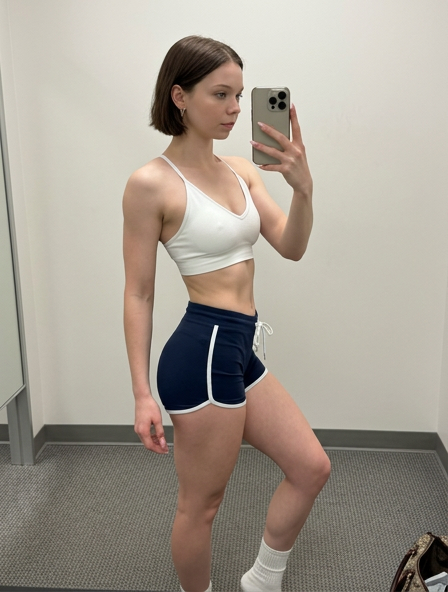 A woman taking a mirror selfie in a fitting room wearing a white sports bra and navy blue athletic shorts against a neutral backdrop.