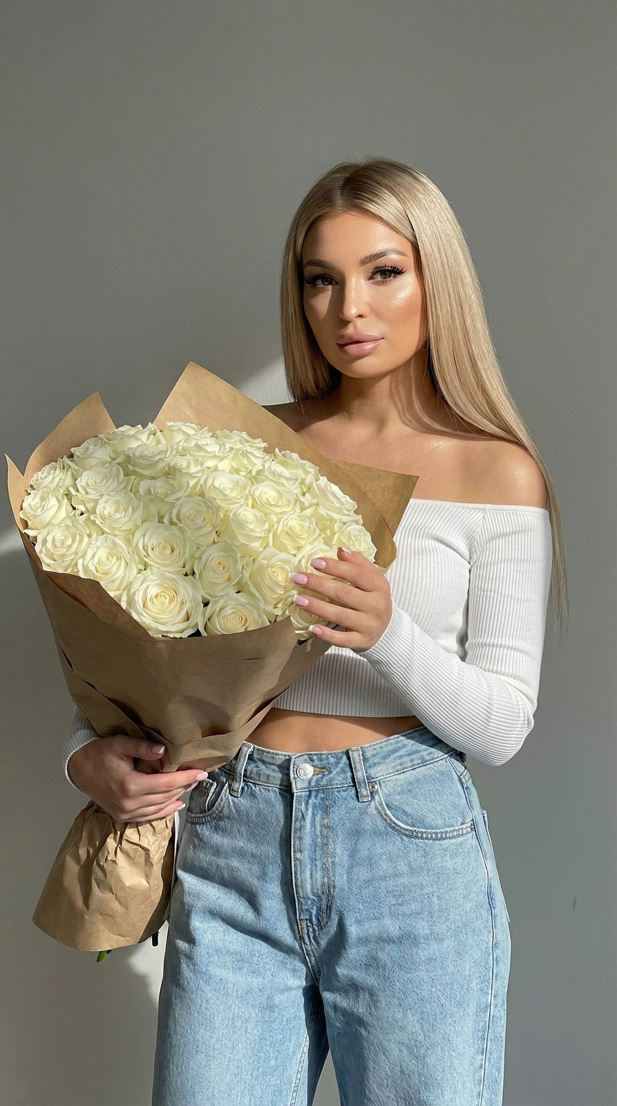 A young woman with long shiny straight hair holding a large bouquet of white roses wrapped in brown kraft paper against a grey wall.