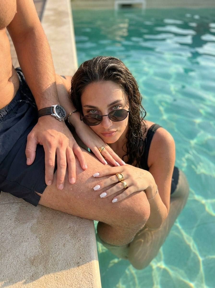 A woman in a swimsuit with wet hair, sunglasses, and accessories, sitting by the edge of a turquoise pool, capturing the sun's reflection.