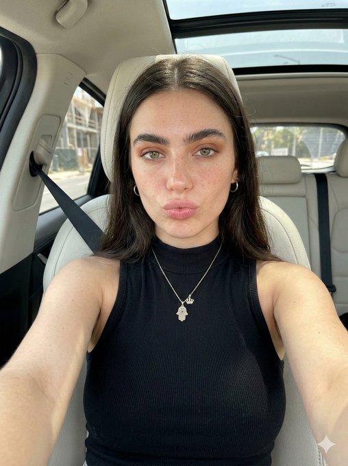 A person with pursed lips in a car selfie, wearing a black sleeveless turtleneck and a gold necklace, with daylight streaming through the sunroof.