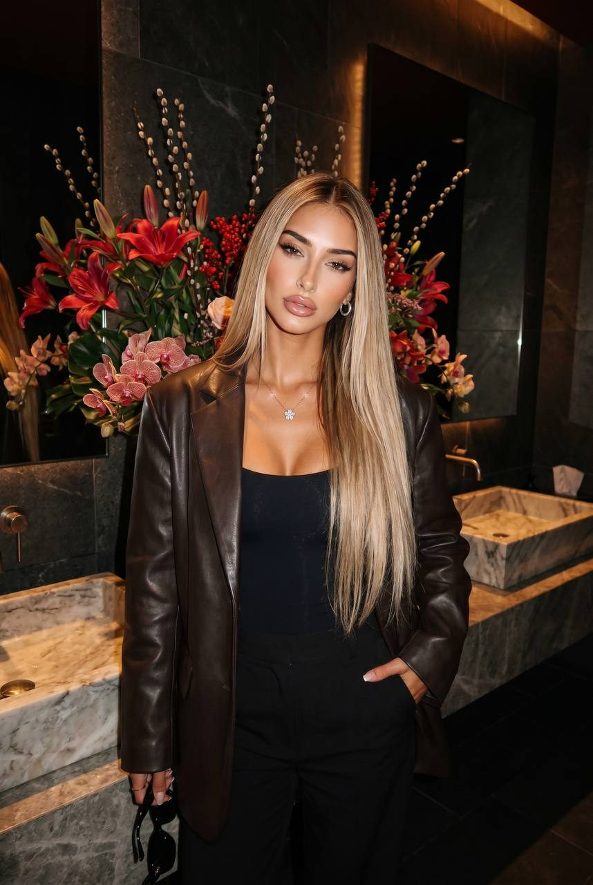 A woman with sleek straight hair in a dark-brown leather blazer standing in a moody luxe restroom, holding sunglasses with a sultry gaze.