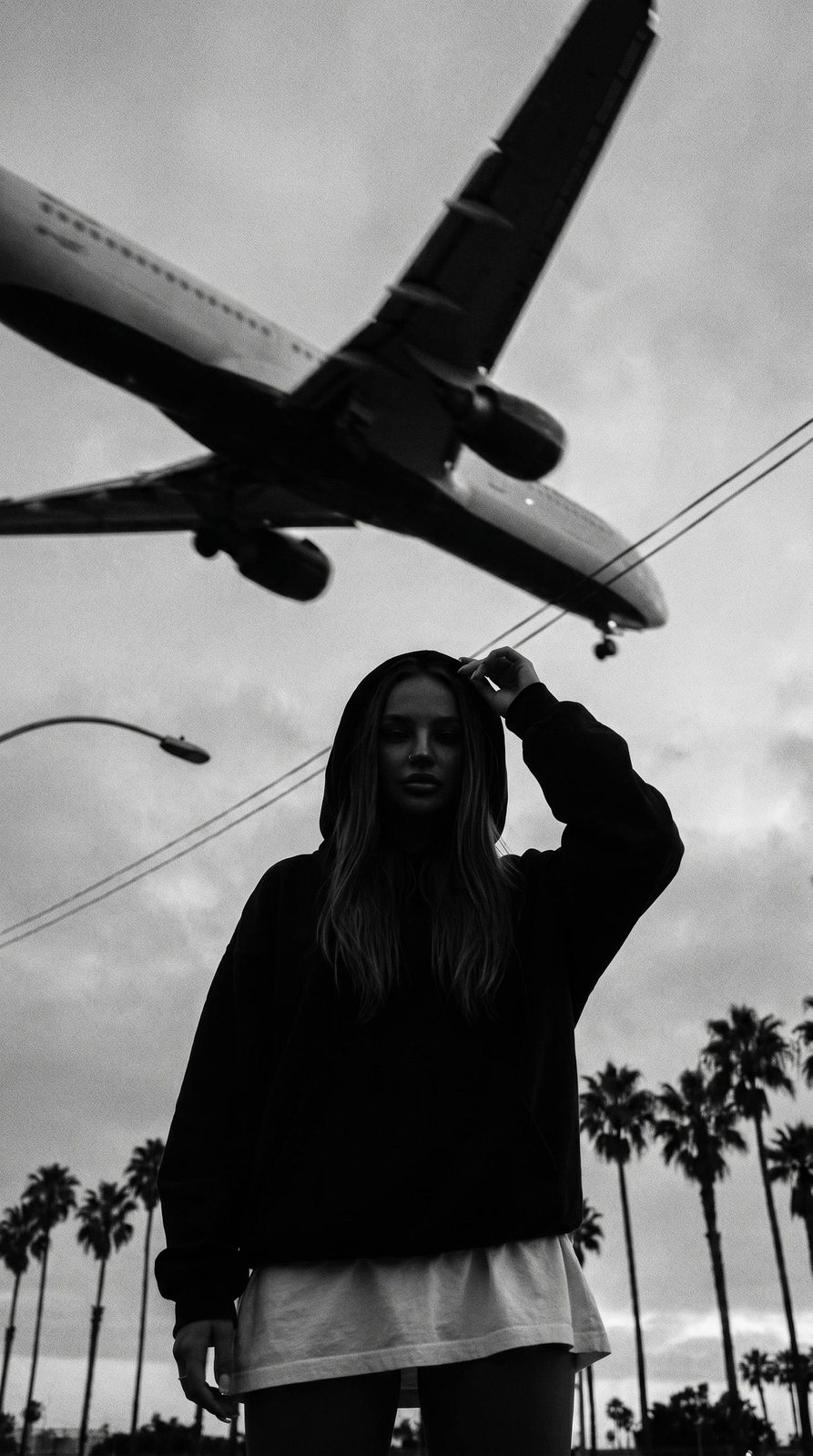 A silhouette of a woman wearing a dark hoodie beneath a low-flying airplane at dusk, framed by palm trees and urban elements.