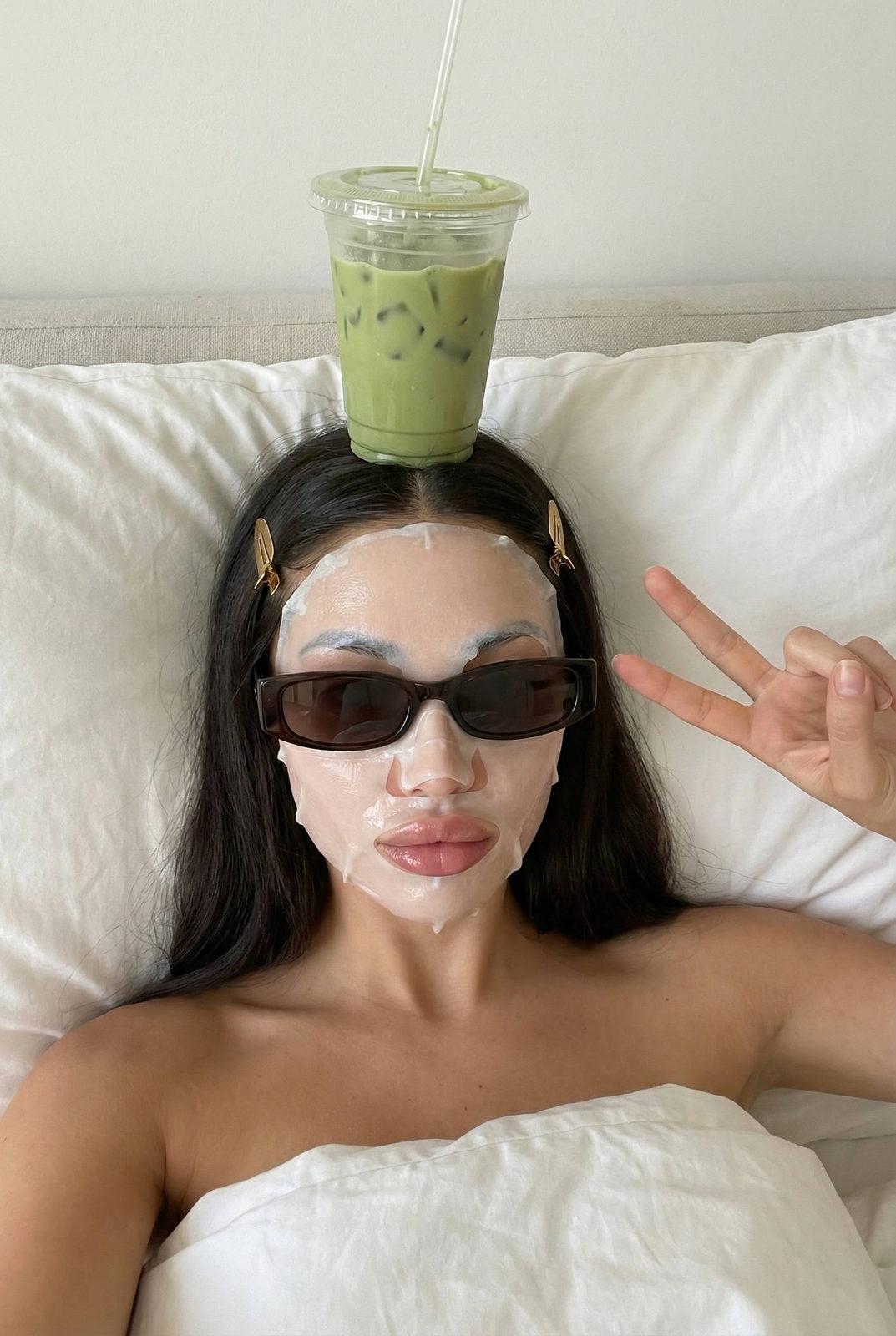 A woman reclines in bed, a cup of iced matcha balanced on her head, forming a peace sign.