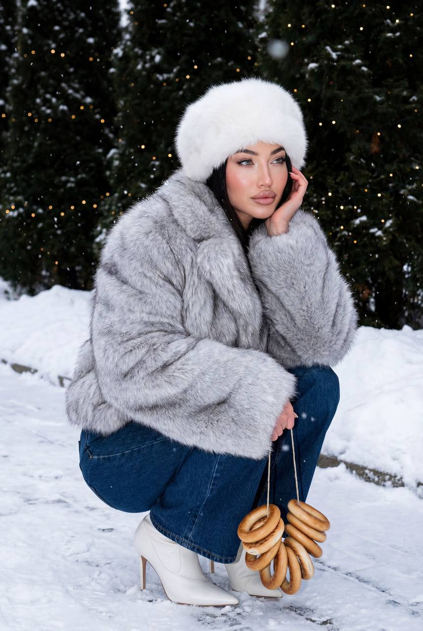 A fashionable woman crouching on a snowy pathway, surrounded by evergreen trees and warm fairy lights. She holds bagels, wearing a silver-gray faux fur coat.
