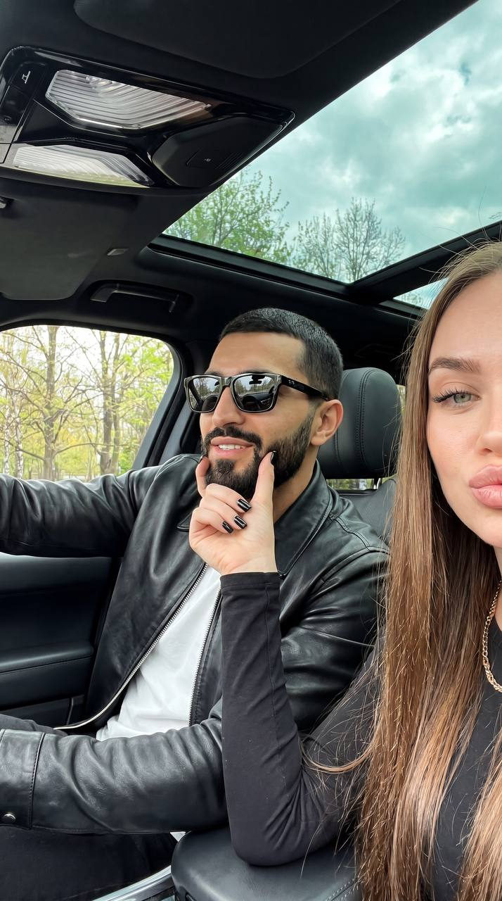 Close-up selfie of two adults in a modern car, showing playful expressions with reflections on sunglasses.