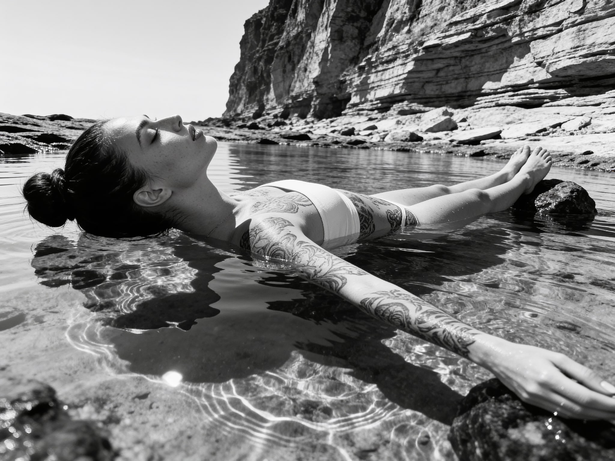 Full-body black and white photograph of a person lying supine in a tide pool, showcasing intricate arm tattoos against a rocky backdrop.